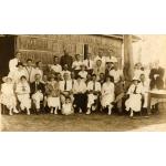 A photo of a group of men and women, with one child, in front of a thatched building. Most of the people are wearing white shirts. 