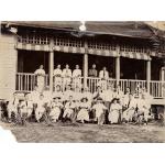 A photo of a group of men and three women, all wearing white. The top row stands on the deck of a building, against a railing. The middle row stands on the ground. The front row is seated in chairs, with spiky plants in front of them.  