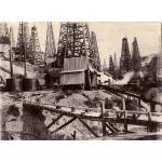 A photo of a group of wooden oil derricks. There is a narrow bridge in the foreground supporting a pipe. There is a cylindrical storage tank on the left with steam boiler stacks sticking up behind it.