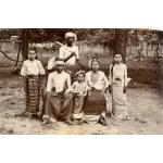 A photo of six Burmese people in traditional clothing. Three are sitting on a wicker chair, and there is a girls standing on either side. A man stands behind holding a machete.