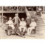 A photo of five men wearing pith helmets, two standing and three sitting in chairs, behind a large cat skin and two sets of animal horns. There are two people and a dog on a set of stairs leading up to a building behind the men.