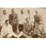 A photo of nine men. Five are sitting on a bench and four are sitting cross-legged on the floor. They are wearing a mixture of light and dark jackets.