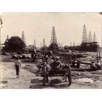 A photo of a group of men with oil derricks in the background. Some men are sitting on carts while others stand around a pile of casing. There is a set of large wagon wheels in the middle of the photo. 