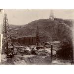 A photo of two metal oil rigs and a third rig up on a hill in behind. There are more hills in the background.