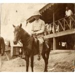 Une photo d’un foreur international vêtu de blanc, coiffé d’un casque colonial et assis sur un cheval. Il est devant un bâtiment monté sur pilotis.  