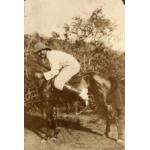 A photo of a man crouched over on a small dark horse. He is wearing a white suit, pants, and shoes as well as a darker hat. There are trees in the background.