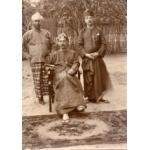 A local man and two International Drillers in front of a fence and trees, with a rug at their feet. They are wearing loose jackets and long pieces of cloth tied like a skirt as well as thong sandals.