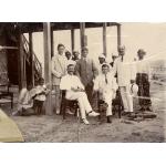 Five International Drillers pose under the stilts which support a house. They are wearing light-coloured suits. Seven local people stand behind them.