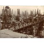 A field full of four-poled oil derricks behind a bridge spanning a low point in the field. There are buildings behind the first row of derricks.