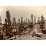 Close to thirty oil derricks with four poles each. There is a bridge leading across a low point in the oil field and several men are walking along the road. There are buildings behind the front row of derricks and a large groups of people are gathered near a pile of casing.