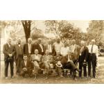 A photo of a group of International Drillers in front of a tree. They are wearing suits. Ten are standing and five are sitting on a bench.