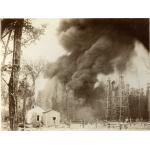 A photo of a column of dark smoke behind two buildings. There is a group of oil derricks on the right.  