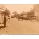  A dirt road with buildings and fences on either side. There are people walking on the street and there is a covered cart, as well. 