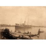 A ship with two masts and a smoke stack floating in the water. There are buildings and trees on the land behind it. In the foreground there is a group of people at a dock with a number of small boats. 