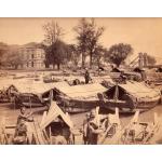 A photo of a group of boats with covers made from reeds beside the shore. There is a large building with windows and arched doorways in the background, with a group of trees.  