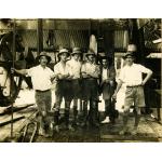 A photo of six International Drillers standing inside an oil rig with metal siding. They are wearing shorts, high socks, and white t-shirts.