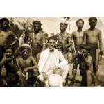 An International Driller wearing a white suit and holding a pith helmet. He is sitting with a group of Dayak men who have a piece of fabric wrapped around their waists. Three of the Dayak men are holding rifles.