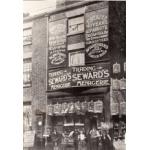 A brick building with birdcages hanging from the top of the windows and along ropes strung across to the next building. There is a group of people outside. Signs on the store front advertise "Trading - Seward's Menagerie" and the animals they sell. 