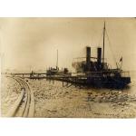 A photo of a tanker tied to a dock along a rocky shoreline. There are four pipes running along the left side of the image and there are boats out in the water in the background. 