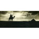 A photo of a man riding a camel and holding a cane. There are two pyramids to the right. The sky behind is full of clouds and the sun in shining down, casting the man and pyramids in shadow.