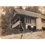 A photo of a bungalow with a thatched roof and sides built on stilts. A dog and a woman sit on the steps going up to the deck, and a man stands to the side of the steps. 