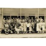 A group of International Drillers and their families outside of a building with latticework on the porch. The men are wearing suits and the women are wearing dresses. 