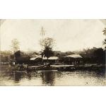 Un groupe de maisons basses à toit de chaume sur les rives d’une rivière, avec des palmiers en arrière-plan. Des bateaux couverts sont ancrés au bord de l’eau.