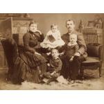 A woman and man sitting with three children between them. They are wearing dark clothing except for the youngest child in the middle who is wearing white. There is a mantle behind them and a fur rug at their feet. 