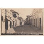 The front of a postcard showing a street with cars and a team of horses in the road.