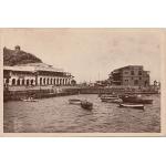 The front of a postcard showing small fishing boats along the edge of the water and two large buildings behind them on land. 