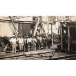 Two International Drillers wearing white and eight labourers in work clothes standing in front of a rig. There is a pile of casing in front of them.  