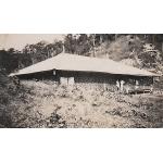 La maison de plain-pied est sur une colline, avec un toit léger et un treillis l’entourant. Trois personnes se tiennent debout dans les escaliers et une autre à quelques mètres.  