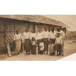 A photo of seven women and four children standing in front of a thatched building. The women are wearing skirts and white shirts. 