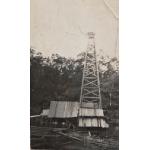 A photo of a covered oil rig. There is a group of men in front of it. There is casing in the foreground and trees in the background. 