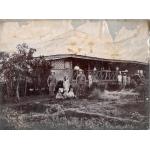 Une photo d’une maison de plain-pied en chaume. Un groupe est à l’extérieur de la maison et deux des hommes portent un uniforme. Il y a des arbres des deux côtés de la photo. 