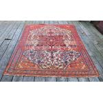 A rectangular red rug with a blue, white, orange, green, and yellow motif in the centre. It is sitting on a wooden deck. 