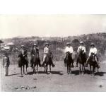 Six International Drillers on horseback. They are wearing pith helmets. A Burmese man stands to their left. There are trees and bushes in the background.