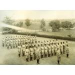 A photo of two rows of police and two rows of rangers in uniform There are three men holding sabres in front of them, ahead of the rows. A fence and line of bushes is in the background.