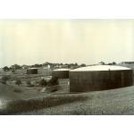 A photo of three oil storage tanks in front of a number of buildings. The sides of the tanks are made from black panels and the roofs are white.