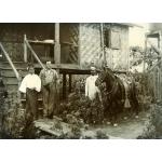 Harry Phillips stands between a Burmese woman and man in front of the steps of a house on stilts. He is holding a rifle and  wearing suspenders. The woman is wearing a long skirt. The man is holding onto a dark horse.