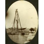 A photo of three men on a wooden tower wit four poles and ropes leading down to a dock. A boat with its mast down is anchored in the water behind them. There is a fourth man on the right beside a pile of objects wrapped in dark material.