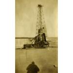 Une tour de forage pour le pétrole à demi recouverte d’un revêtement métallique. De l’équipement mécanique se trouve à la droite. Le sol est plat et en terre et il y a une colline en arrière-plan. On aperçoit l’ombre du photographe à l’avant de la photo