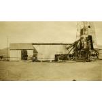 An oil derrick covered in metal siding. There is mechanical equipment on the right. The ground is flat dirt and there is a hill in the background.