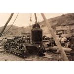 A photo of a partially assembled bulldozer beneath a wooden tripod. There is one man at the front of the bulldozer and four behind it. There are hills in the background. 