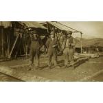 Three International Drillers wearing work clothes and hats, standing in front of an oil derrick with a roof made from metal sheeting. There are dirt hills in the background.