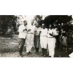 A photo of five men wearing white shirts stand together, holding small glasses of alcohol. There is a group of people behind them to the right.