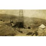 A metal oil rig. There are men standing around it and near a car. There are metal cylinders in the foreground and a tent on either side of the rig. There is a cylindrical steam boiler behind the rig. There are hills in the background.