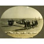 A photo of a group of men carrying a pipe. The pipe is attached to two beams and the men are standing at the front and back of the pipe and supporting the beam on their backs. The ground is flat and there is a line of white rocks in the foreground.