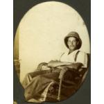 A man sitting in a wicker chair with his arms crossed and legs extended. He is wearing a pith helmet, white t-shirt, dark pants, and suspenders.