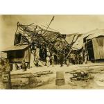 A photo of a metal derrick that has fallen over on top of a structure with a metal roof and siding. A group of people are standing in front of it looking at the damage. There are metal cylinders and casing in the foreground.